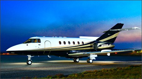 A private jet is parked on a runway at dusk, with its lights on and the sky transitioning from blue to orange.