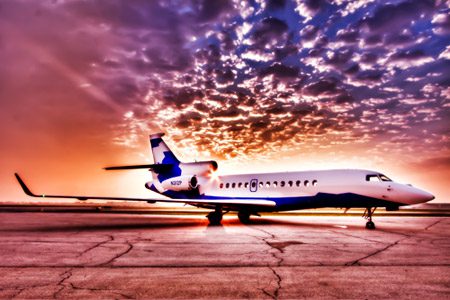A white and blue private jet is parked on an airport tarmac at sunset, with dramatic clouds and orange sky in the background.