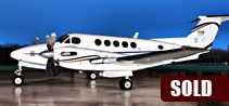 A white King Air B200 twin-turboprop airplane is parked on a wet tarmac at dusk, with a red "SOLD" label in the lower right corner.