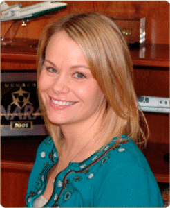 A woman with straight blonde hair and a teal embroidered top smiles at the camera, standing in front of a wooden shelf with model airplanes and awards.