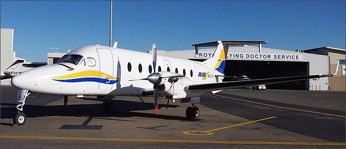 A 1995 Beechcraft 1900D For Sale UE-0143 by General Aviation Services, featuring yellow and blue accents, is parked on an airport tarmac in front of a building labeled "Royal Flying Doctor Service.