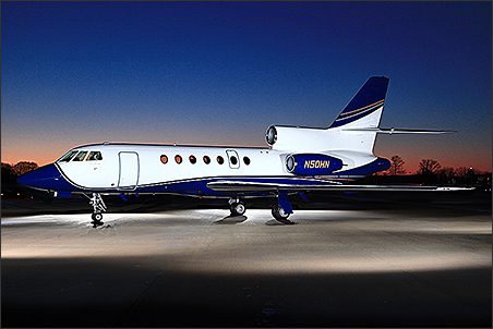A Dassault Falcon 50 for sale, this used corporate jet aircraft is parked on a runway at dusk, illuminated by lights, with a dark blue sky in the background.