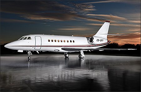 A Dassault Falcon 2000EX EASy business jet with red and black stripes is parked on a reflective surface at dusk, with trees and a colorful sky in the background—a stunning used 2000 jet aircraft for sale.