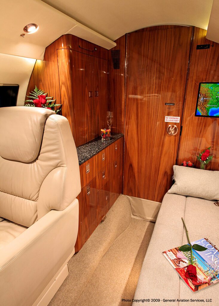 A luxurious private jet interior with wood paneling, leather seating, a small countertop with a wine glass, and magazines on a side table.