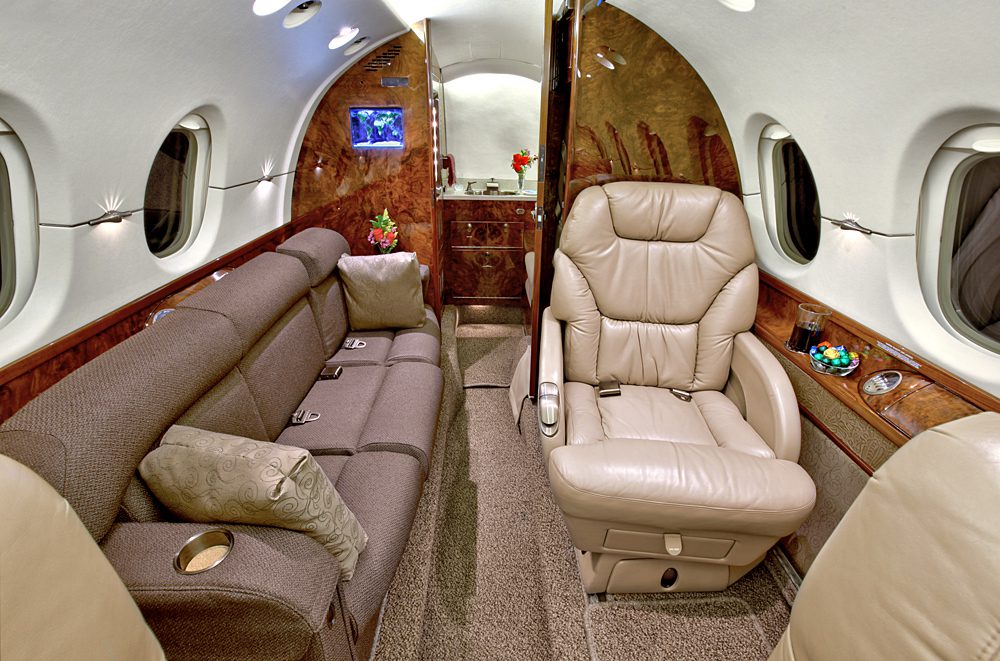 Interior of a private jet with a brown leather armchair, gray sofa, wood paneling, windows, and a small table with a bowl of candy.