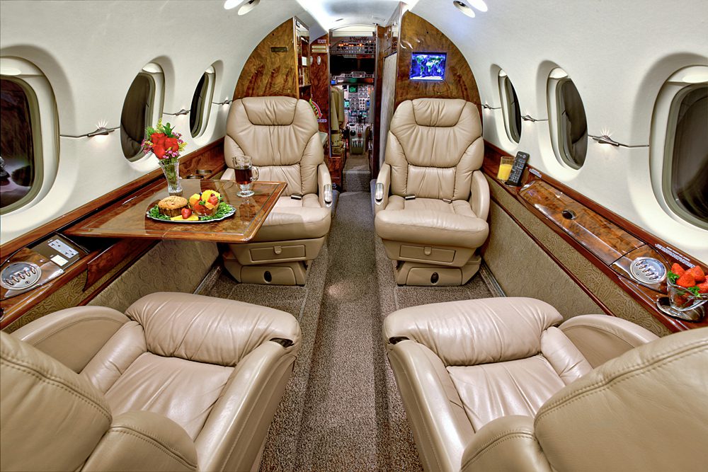 Interior of a private jet with four beige leather seats, a wooden table set with food and drinks, large windows, and a view toward the cockpit.