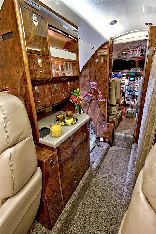 View of a private jet cabin with a wood-paneled galley, snacks and drinks on a counter, and the cockpit visible in the background.
