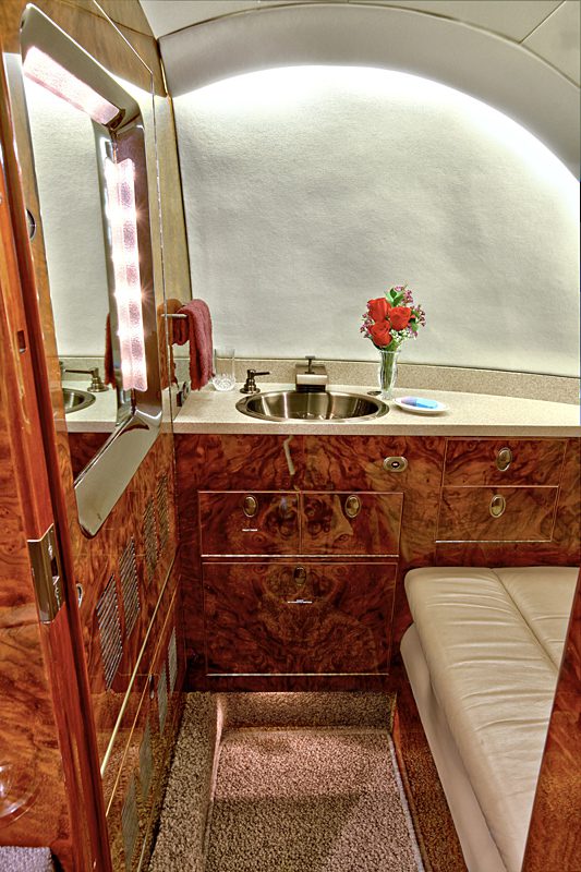 Compact, luxurious private jet bathroom with wood paneling, a cushioned seat, sink, mirror with lights, towel, and a vase of red flowers on the countertop.