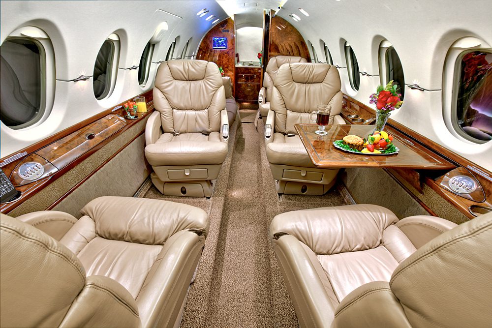 Luxurious private jet cabin with cream leather seats, wood paneling, a table set with wine and food, and windows along both sides.