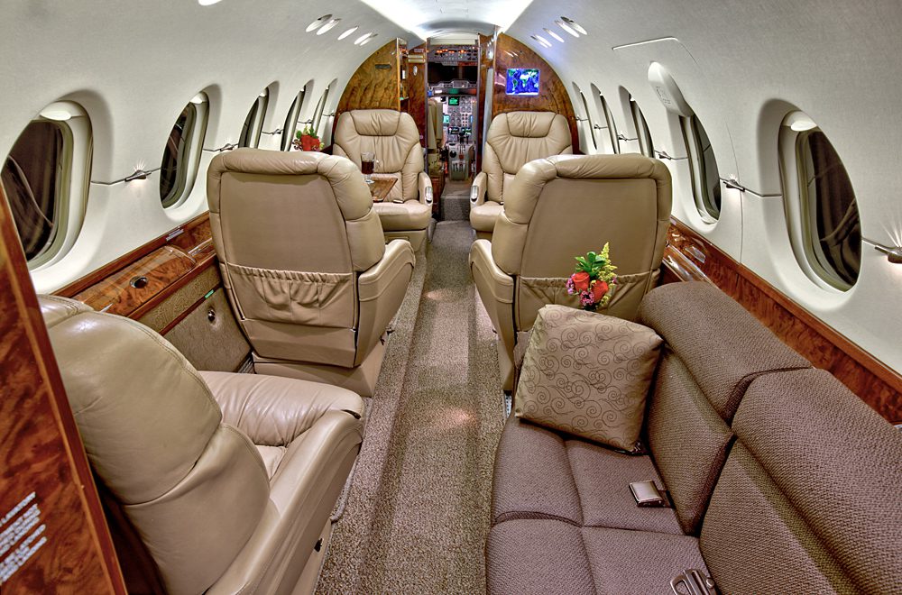 Interior of a private jet featuring beige leather seats, a brown carpet, wood paneling, and flower arrangements on small tables beside the seats.