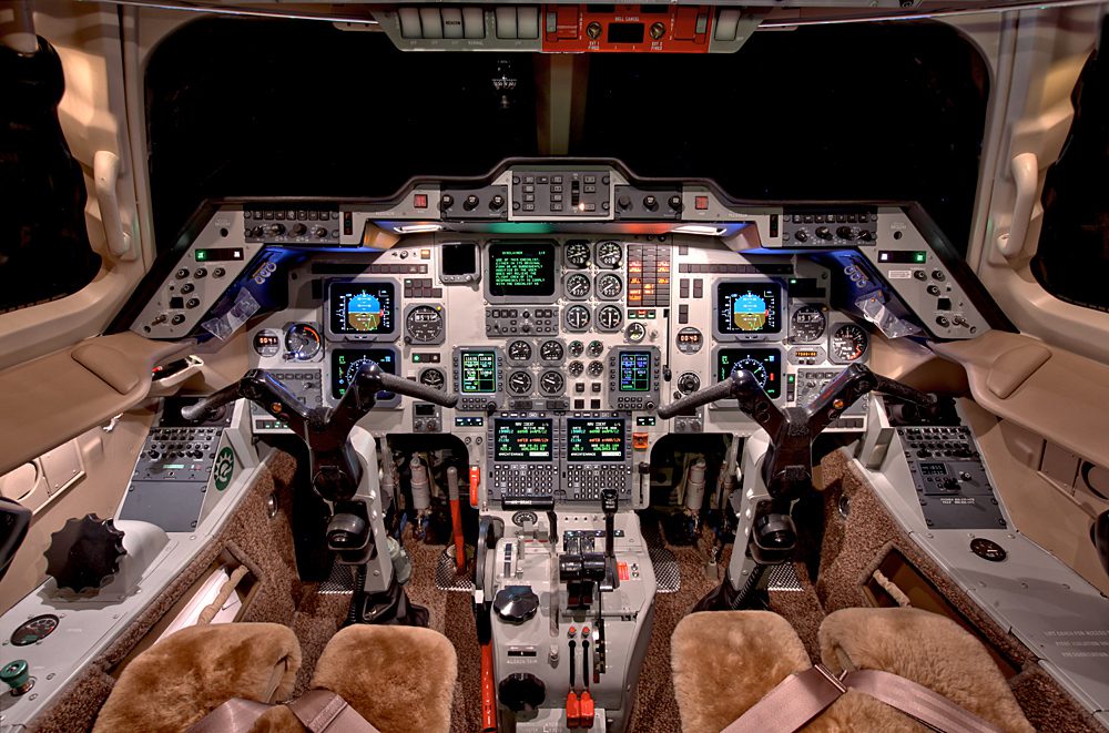 Cockpit interior of an aircraft featuring two pilot seats, control yokes, numerous switches, levers, digital screens, and analog gauges.