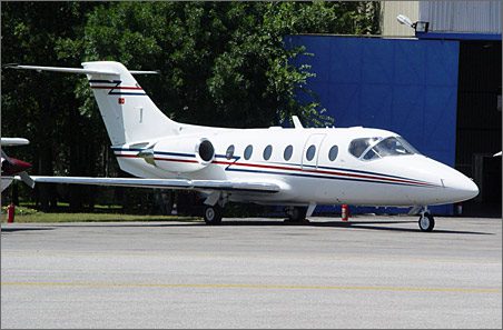 A small white Beechjet 400A with red and blue stripes, ideal for corporate business jet sales, is parked on the tarmac near a blue hangar and trees.
