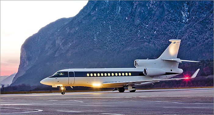 A 2008 Falcon 7x for sale is parked on an airport runway with mountains in the background during sunset, available for private showing at EBACE.