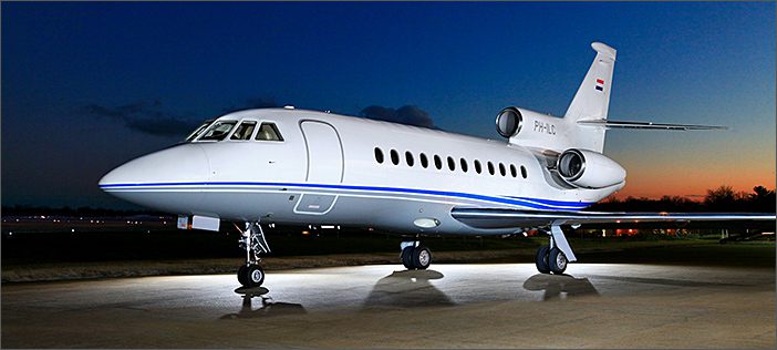A white Dassault Falcon 900B business aircraft with blue accents is parked on a lit tarmac at dusk, showcasing its sleek design against the darkening sky—a prime example of a used corporate jet for sale.