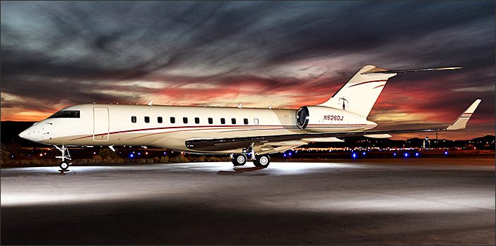 A Bombardier Global 5000 used corporate jet is parked on a runway at dusk, with dramatic, colorful clouds in the sky and runway lights illuminating the aircraft.