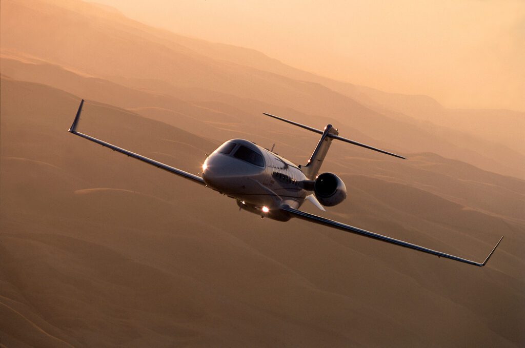 A private jet flies over a desert landscape at sunset, with soft orange light illuminating the scene.