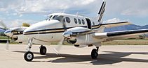 A twin-engine propeller airplane is parked on a tarmac, with mountains and a partly cloudy sky in the background.