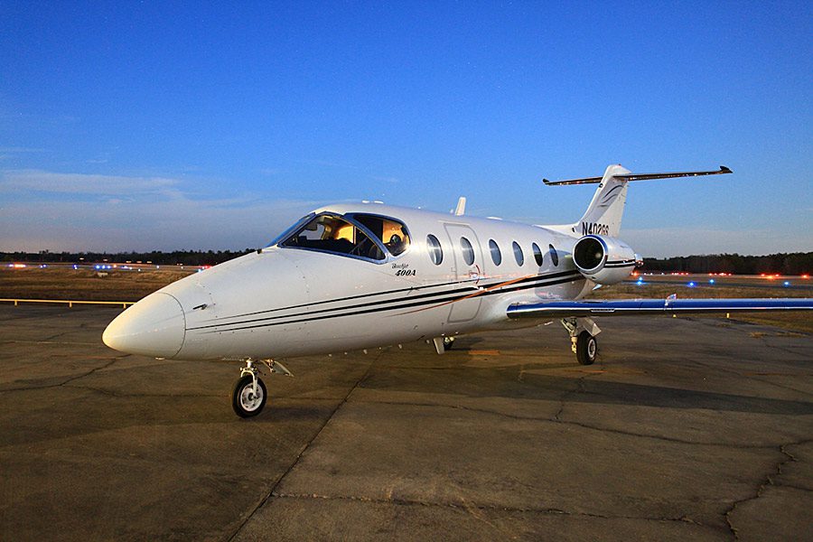 A white private jet with black stripes is parked on an airport tarmac at dusk, with runway lights and a clear sky in the background.