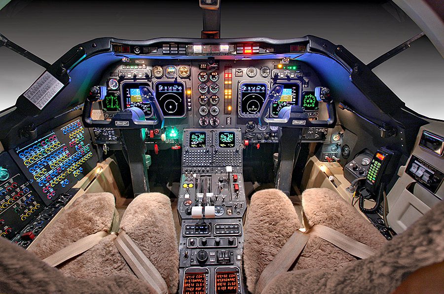Cockpit interior of an aircraft showing two pilot seats, numerous control panels, switches, levers, displays, and illuminated instruments.
