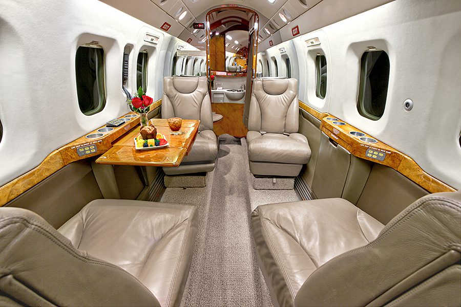 The interior of a private jet featuring four leather seats, a wood table with fruit and flowers, and windows along both sides.