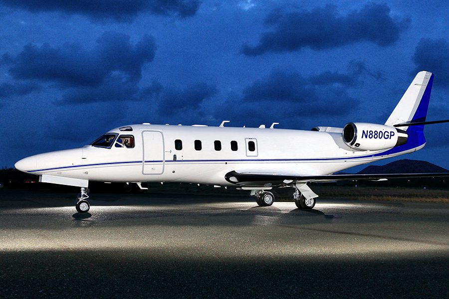 A white private jet with blue accents is parked on a runway at night under a cloudy sky, illuminated by artificial light.
