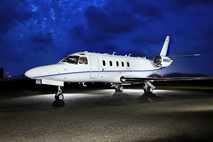 A white private jet is parked on a wet tarmac at night under a dark, cloudy sky, illuminated by lights.