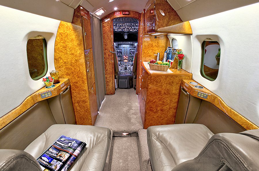 The interior of a private jet with beige leather seats, wood paneling, a small counter with flowers, and a view into the cockpit.