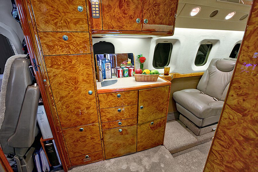Luxurious jet interior featuring a small bar area with drinks, fruit, and snacks, glossy wood cabinets, and a cream leather seat by the windows.