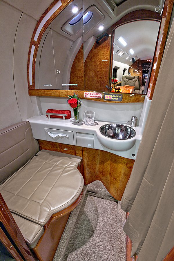 Private jet lavatory with a leather bench seat, small sink, rose in a vase, glass, and mirror, separated by a curtain; cockpit visible in the background.