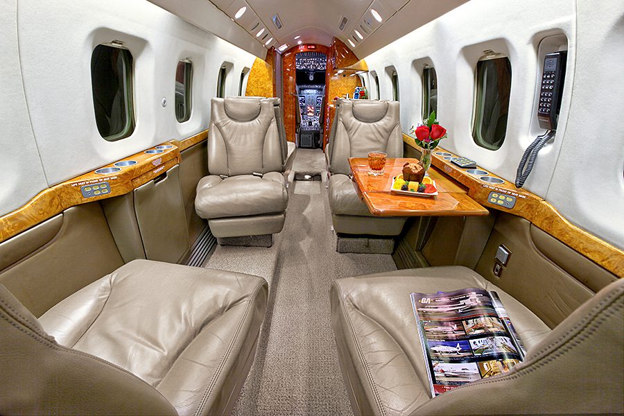 The interior of a private jet with four leather seats, a wooden table holding refreshments and flowers, and a magazine on one seat. The cockpit is visible in the background.