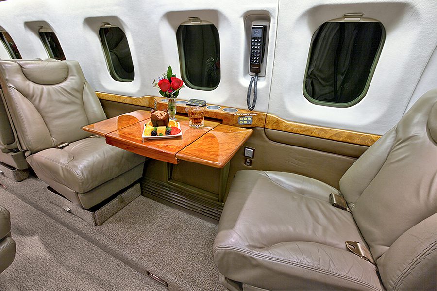 Two leather seats face a wooden table with a drink, dessert, and vase of roses inside a private jet cabin; three windows and a phone are visible on the wall.