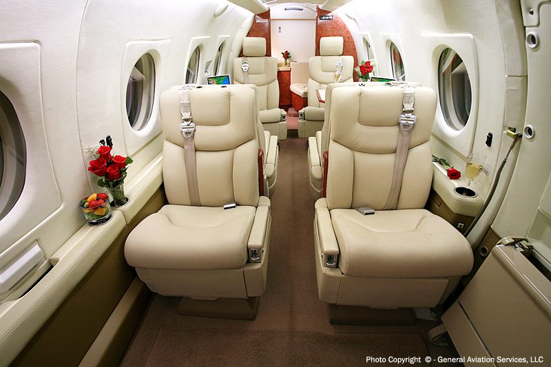Luxurious private jet interior with cream leather seats, small tables, windows on both sides, and flower arrangements on armrests.