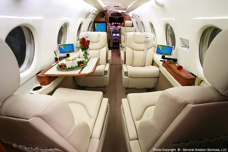 Interior of a private jet with four cream leather seats, a table with a meal and flowers, and screens displaying maps; cockpit is visible in the background.