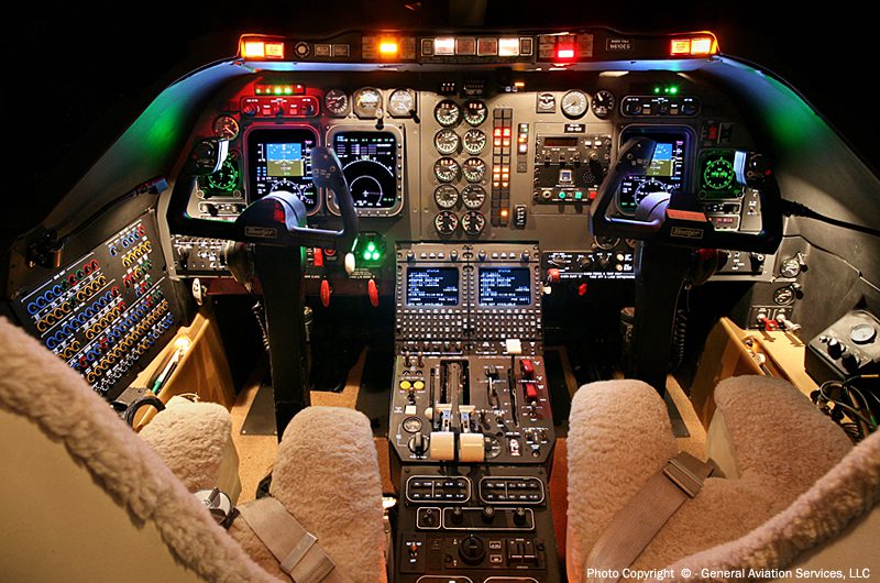 Cockpit interior of an aircraft with illuminated control panels, instrument gauges, and two seats covered in sheepskin.