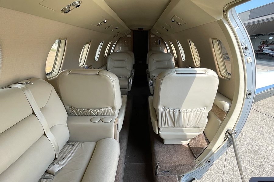 Interior view of a small private jet featuring light-colored leather seats, armrests, and seatbelts, with the aircraft door open to the outside.