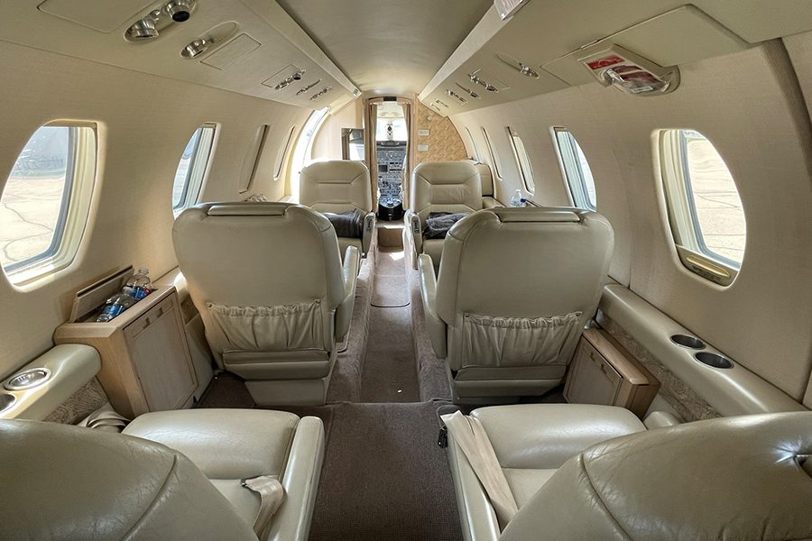 The interior of a small private jet showing six cream-colored leather seats, windows on both sides, and a view toward the cockpit at the front.