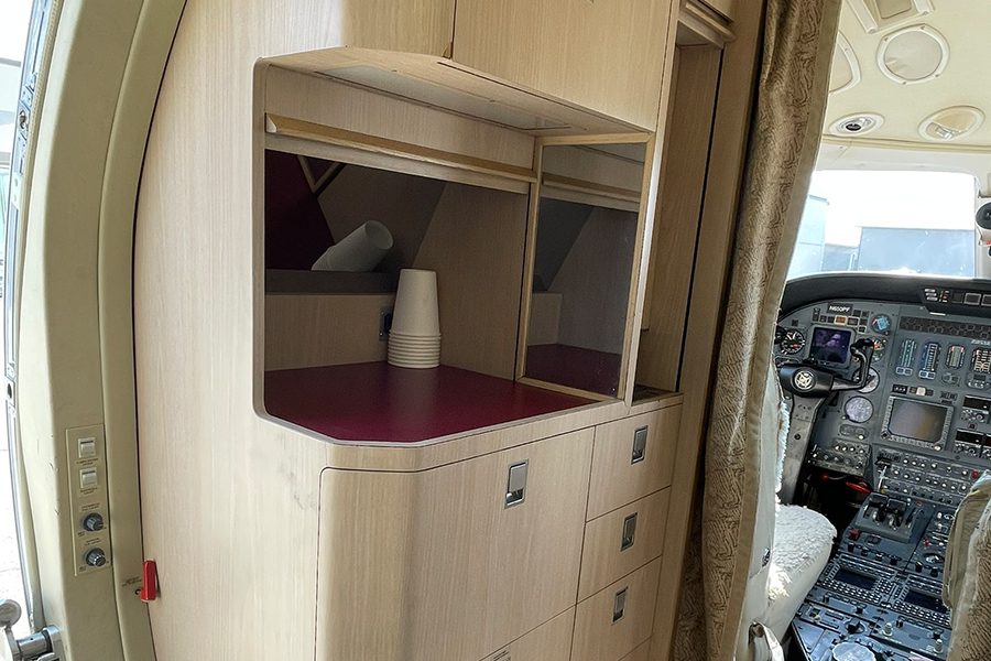 Interior view of a small aircraft showing a kitchenette with stacked cups on a counter and the cockpit with instrument panels visible to the right.