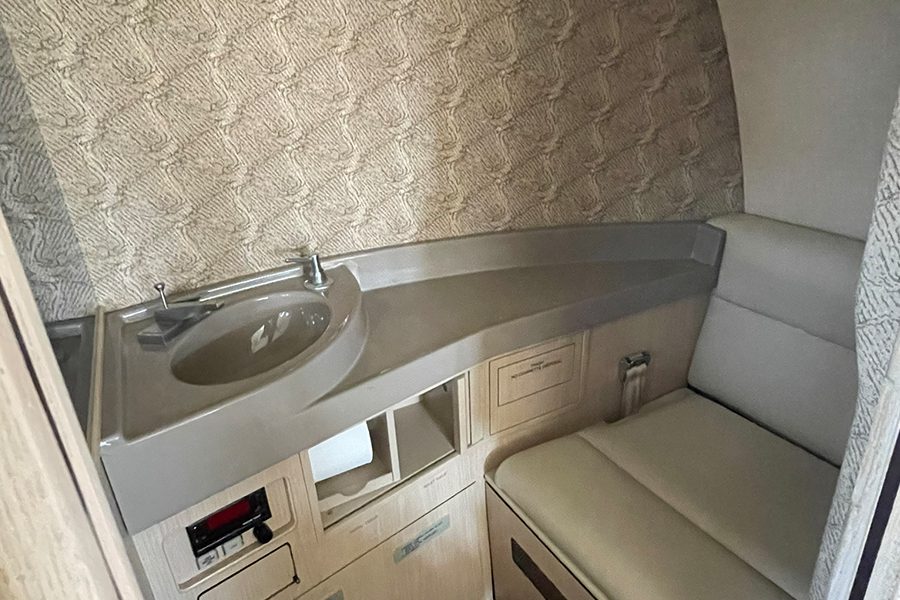 Compact airplane lavatory with a small sink, countertop, storage compartments, and a cushioned bench seat against a patterned wall.