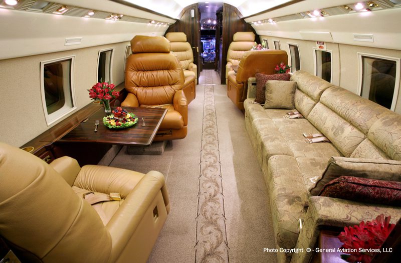 Interior of a private jet featuring leather seats, a long sofa with cushions, tables with floral arrangements, and a view toward the cockpit.