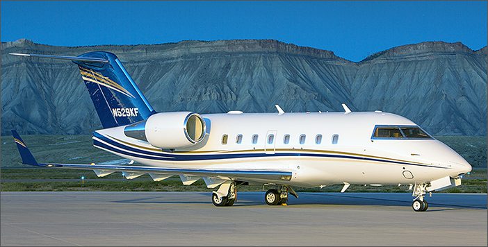 A white 2001 Challenger 604, serial number 5477, with blue and gold accents is parked on a runway against a rocky mountain backdrop—an impressive used corporate jet now for sale.