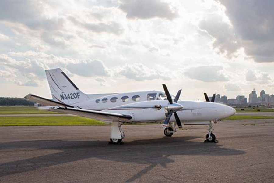 A white twin-engine propeller airplane is parked on an airport tarmac with a city skyline and cloudy sky in the background.