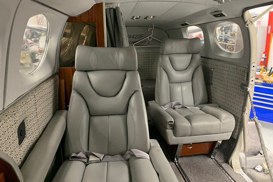 Two gray leather passenger seats inside a small aircraft cabin, with patterned side panels and seatbelts.