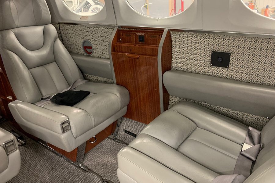 Two gray leather seats with seatbelts in a small private jet cabin, featuring wood paneling and patterned sidewalls.
