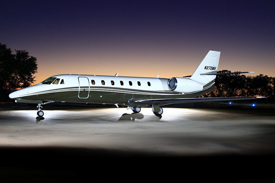 A private jet is parked on a lit tarmac at dusk with trees and a darkening sky in the background.