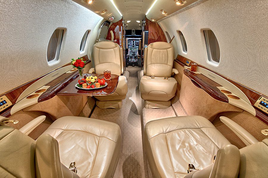 Luxurious private jet interior with four beige leather seats, a table set with fruit and drinks, wood accents, and a cockpit visible in the background.