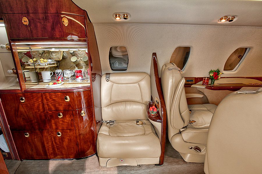 Interior of a private jet with cream leather seats, a wood-paneled cabinet stocked with glassware, and a small table with flowers near the windows.