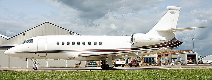 A white Falcon 2000 used business jet is parked on the tarmac near several hangars and industrial buildings under a cloudy sky.