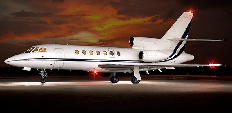 A white private jet is parked on a runway at sunset, with its lights on and a dramatic orange sky in the background.