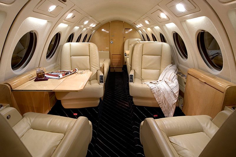 Interior of a private jet featuring four beige leather seats, fold-out table with drink and magazines, and a blanket on one seat; circular windows line both sides.
