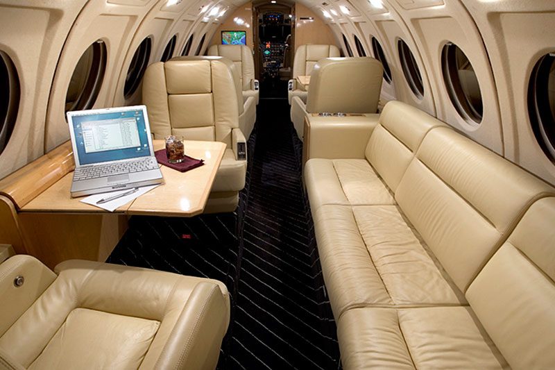 Interior of a private jet with beige leather seats, a fold-out table holding a laptop and a glass, and round windows along the cabin walls.